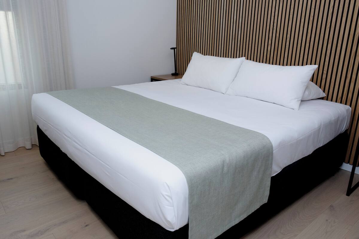 Cama bem arrumada com lençóis brancos e um cobertor verde, cabeceira de madeira e mesa de cabeceira com luminária.