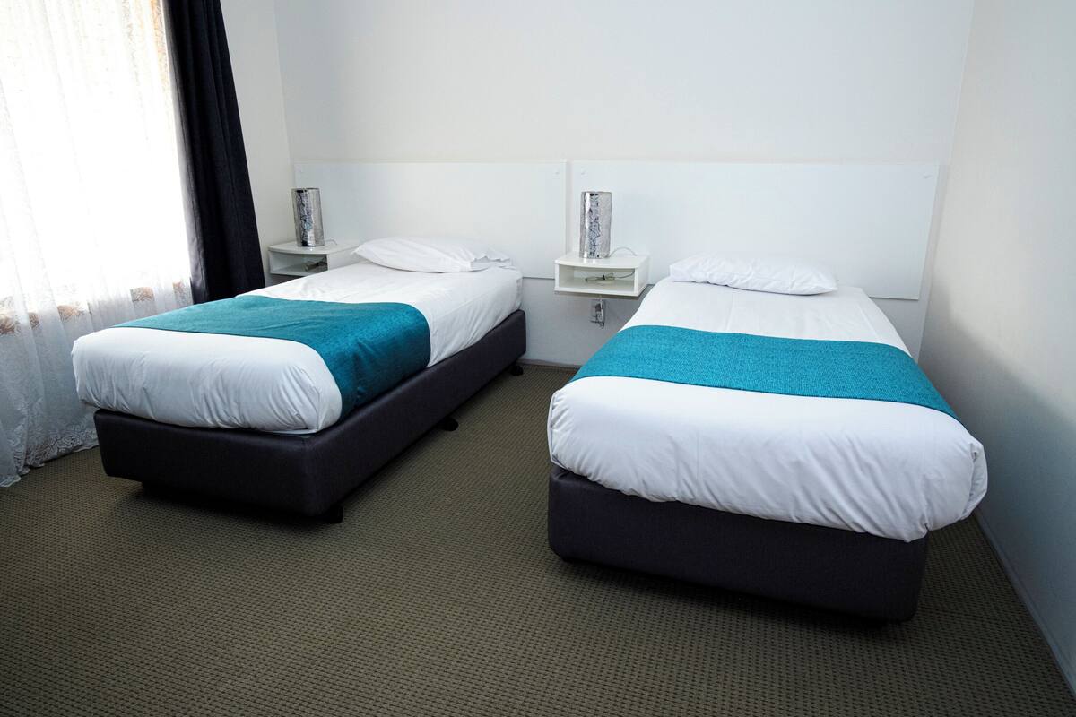 Quarto de hotel com duas camas de solteiro, uma pequena mesa de cabeceira com abajur e uma janela com cortinas.