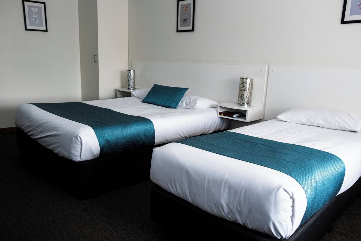 Duas camas de solteiro com roupa de cama branca e azul-turquesa, uma mesinha de cabeceira com abajur, e quadros nas paredes.