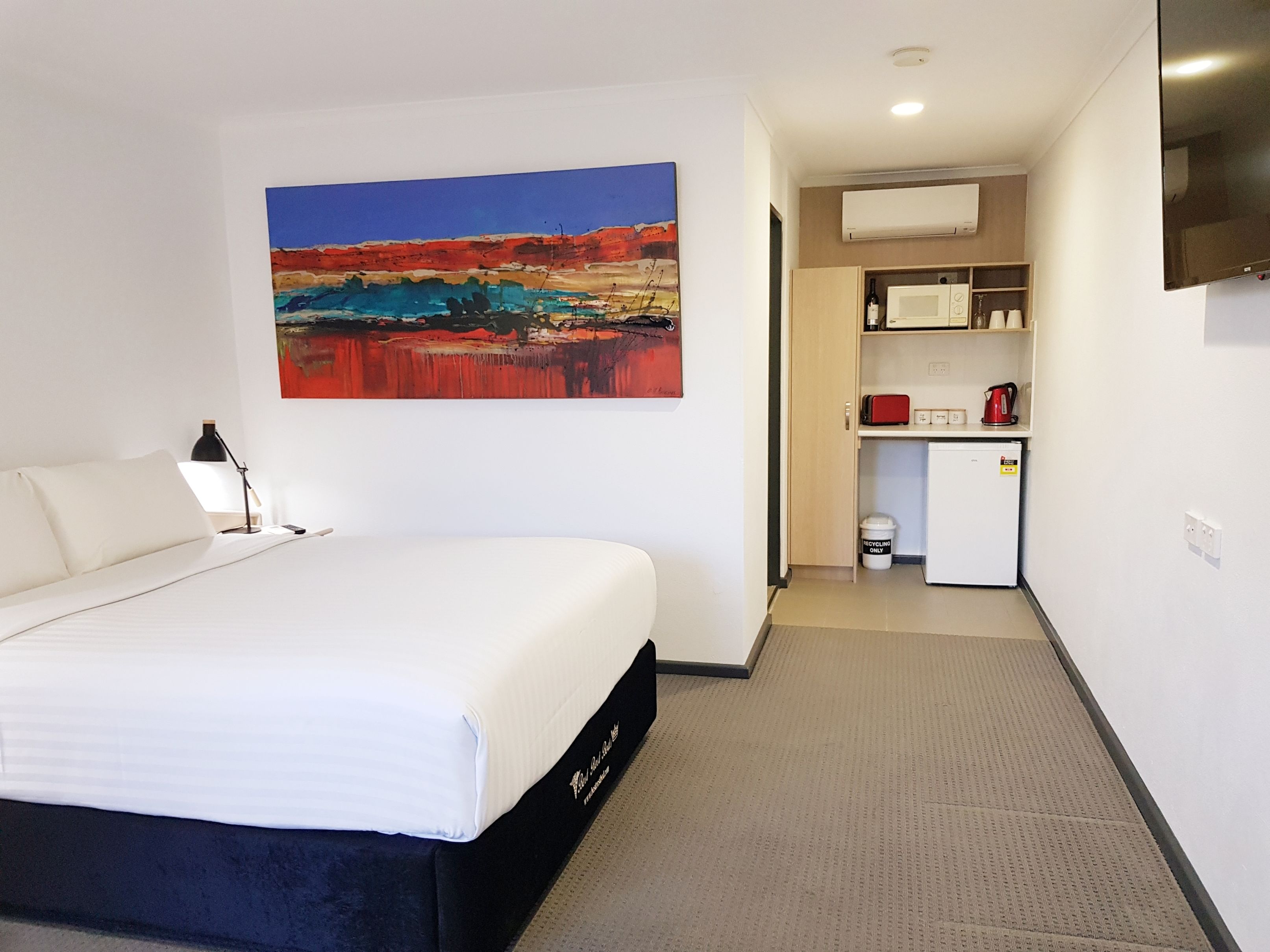 A hotel room with a bed, a painting, a kitchenette, and a TV.