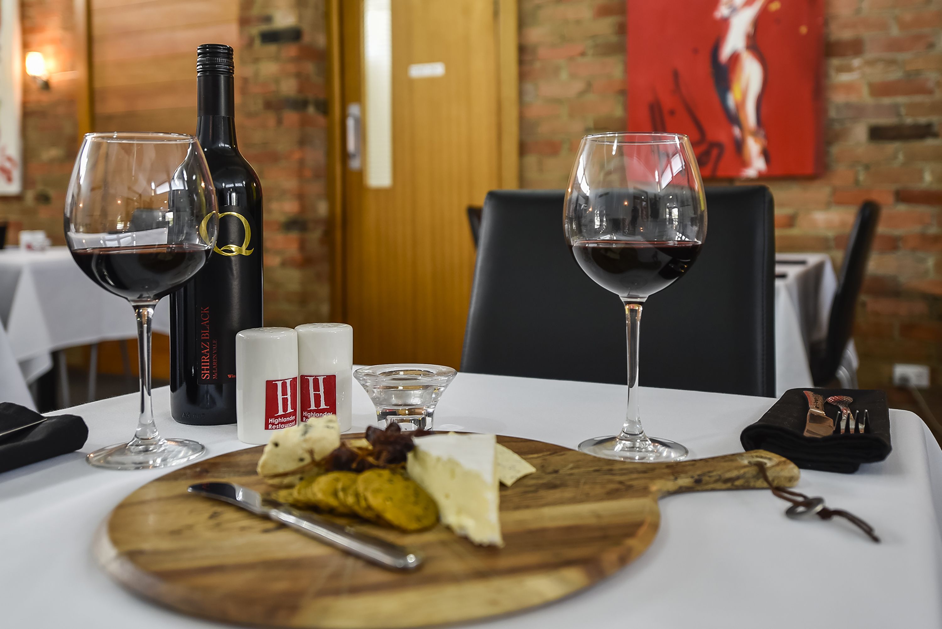 Una mesa dispuesta con copas de vino, una botella de Shiraz Black, queso y una tabla de madera con un cuchillo.