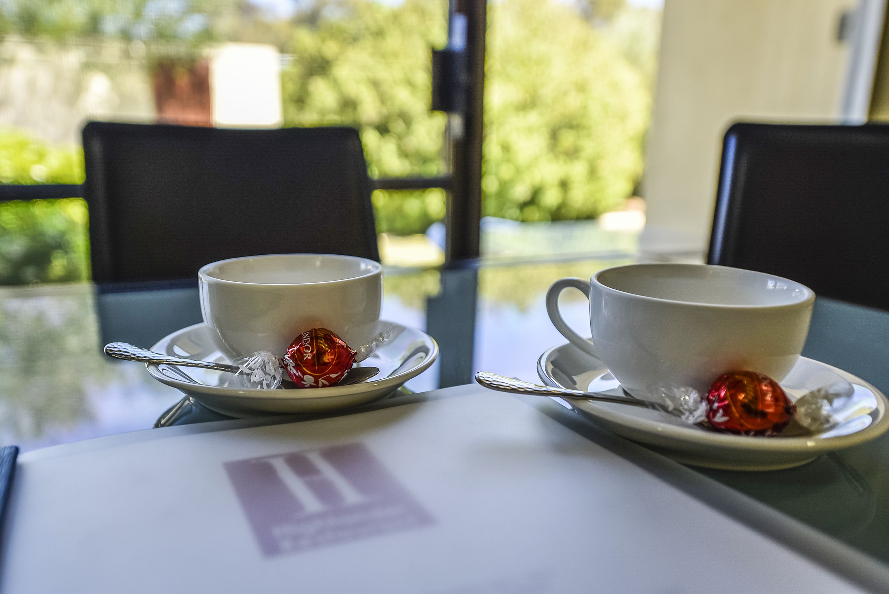 Two white cups with saucers on a glass table, each with a spoon and a wrapped confectionery.