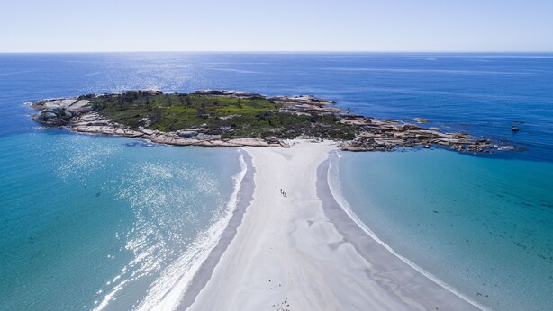 Vue aérienne d’une plage avec une petite île, des eaux d’un bleu limpide et un rivage sablonneux.