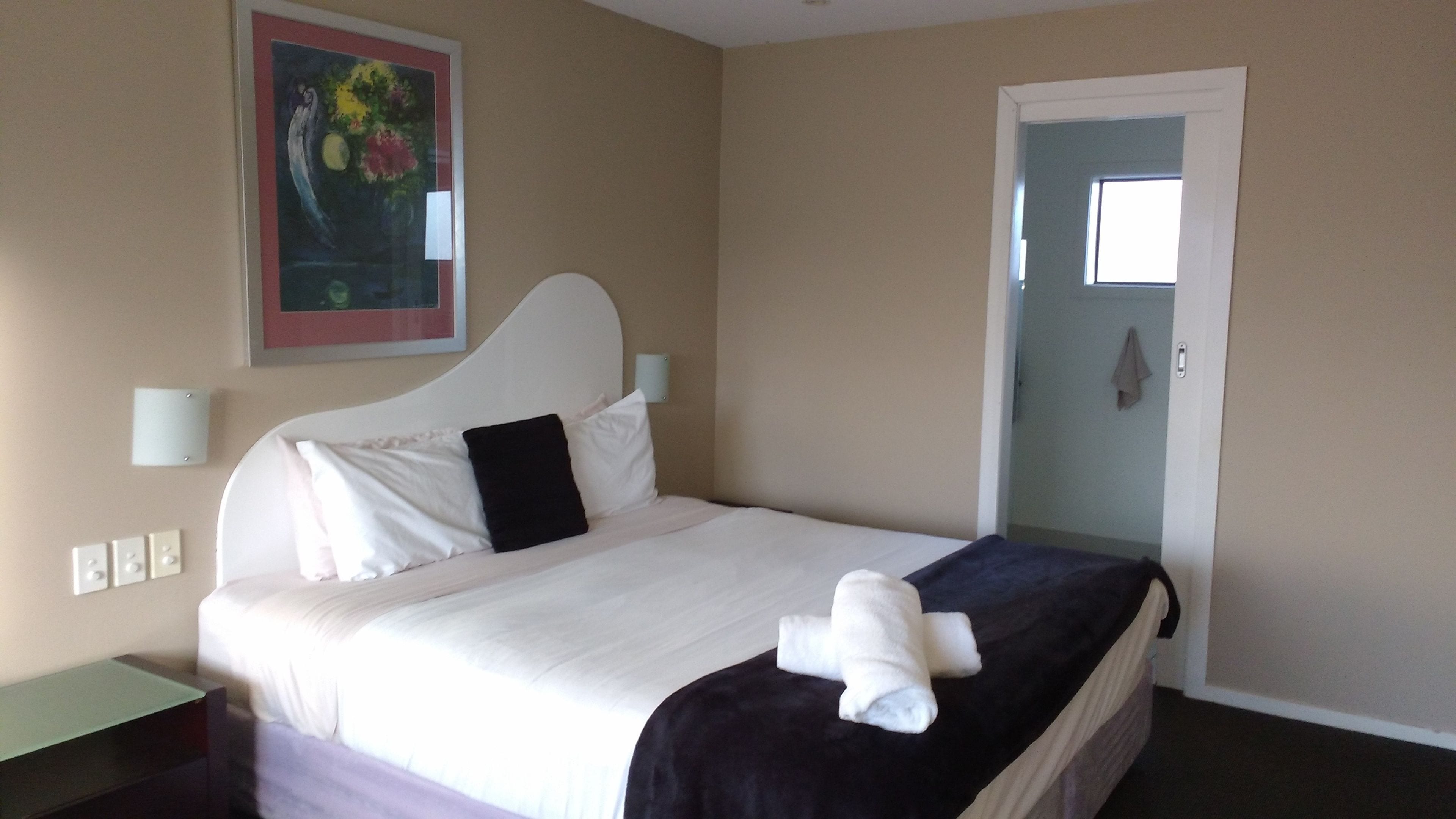 A hotel room with a bed, a painting on the wall, a door, and a nightstand.