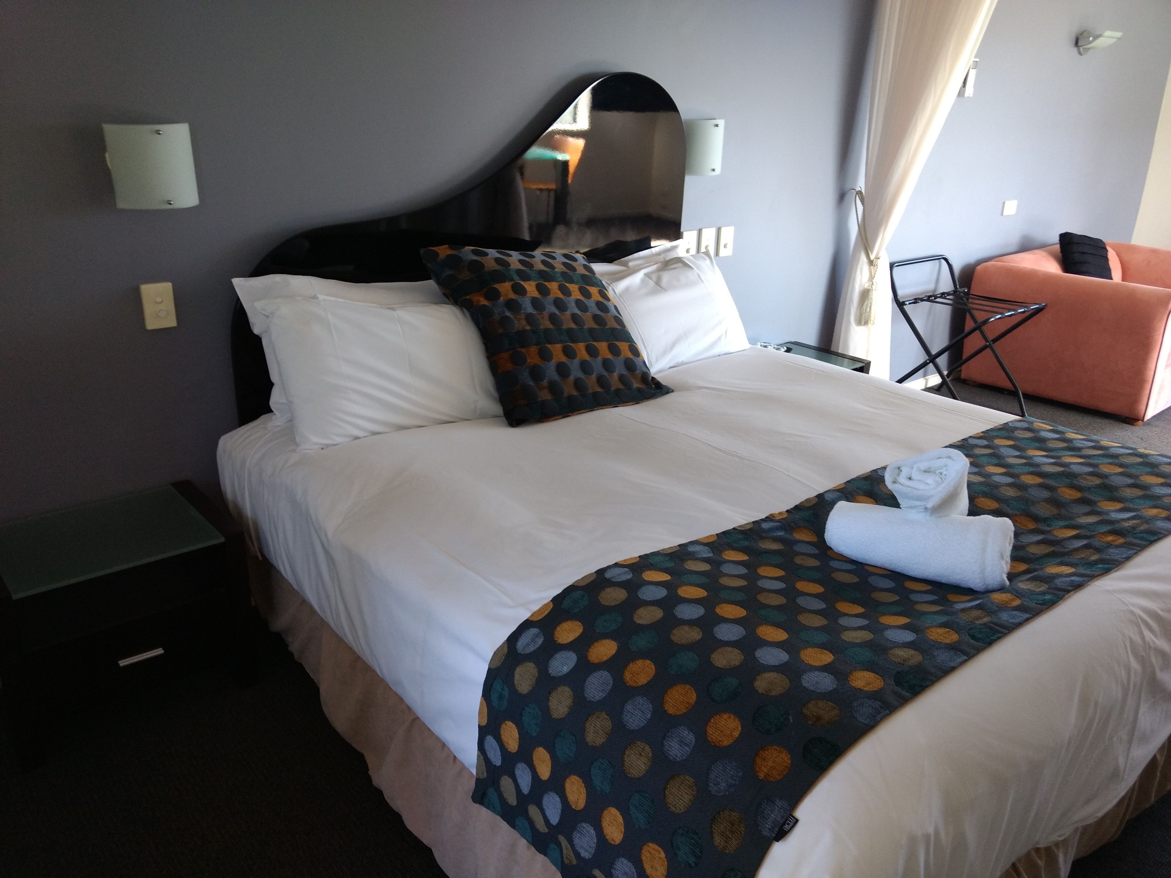 A neatly made bed with white linens and a polka dot blanket, a small bedside table, and a chair with a towel on it.