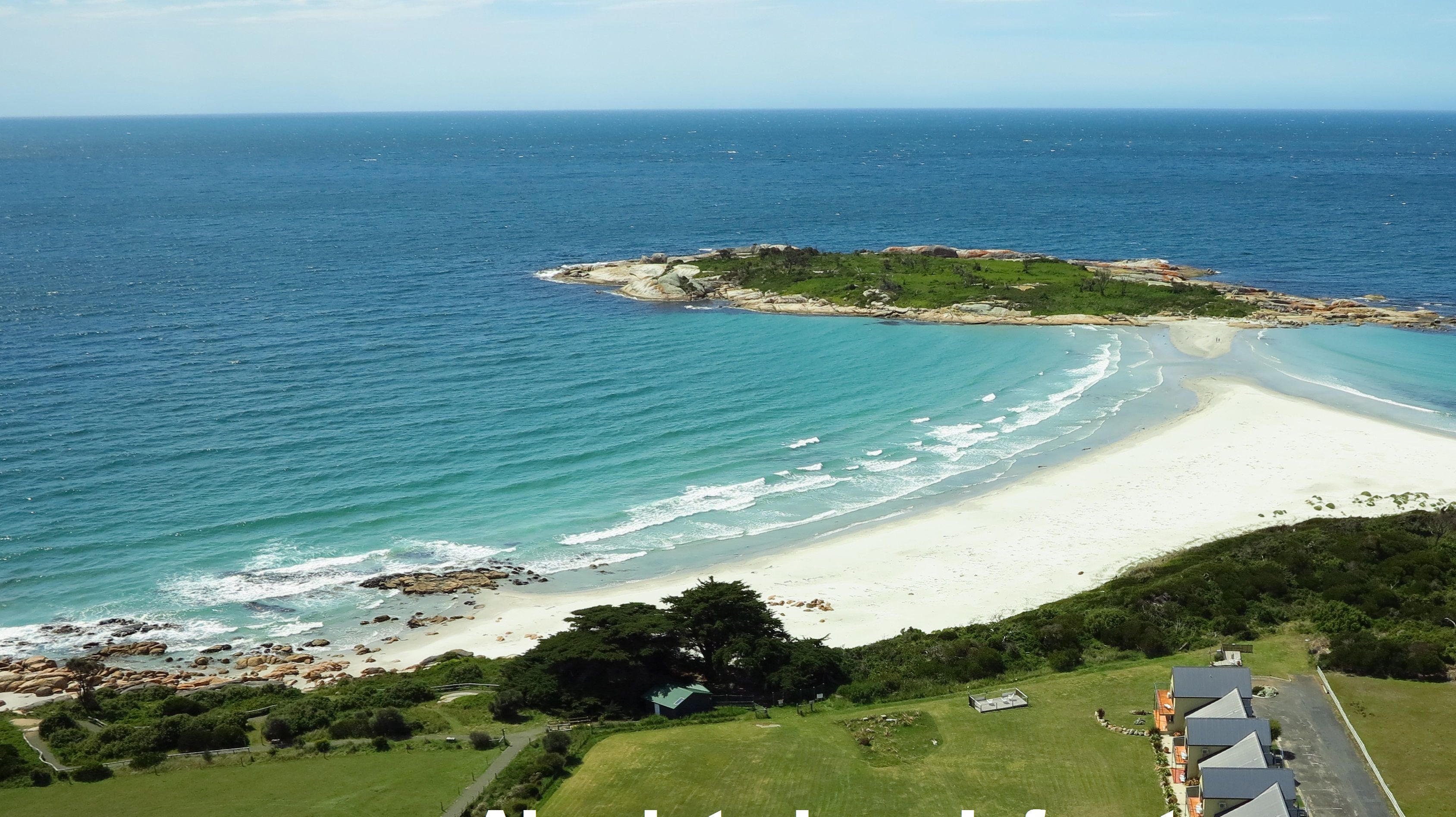 A coastal landscape with a sandy beach, rocky outcrops, and a small green area with a building.