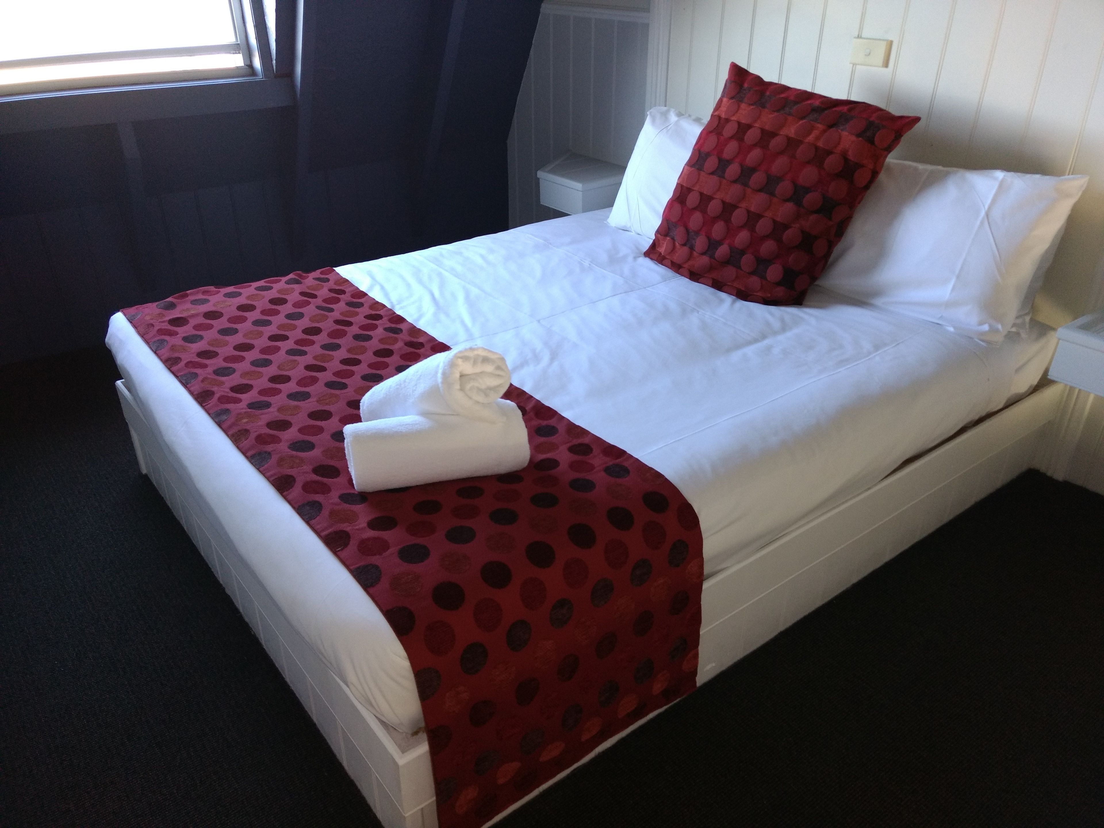Una cama bien hecha con sábanas blancas, una colcha roja con lunares y una almohada roja y negra. Hay dos toallas blancas dobladas sobre la cama.
