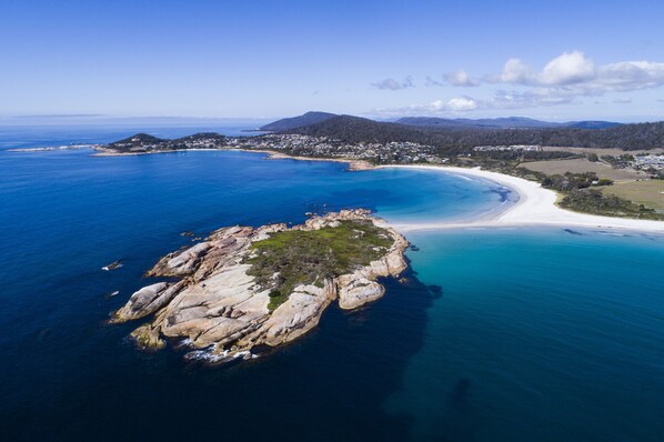 An aerial view of a coastal area with a prominent rocky outcrop surrounded by clear blue waters, a sandy beach, and a residential area with houses and buildings in the background. - Diamond Island Resort & Penguin viewing (Bicheno)