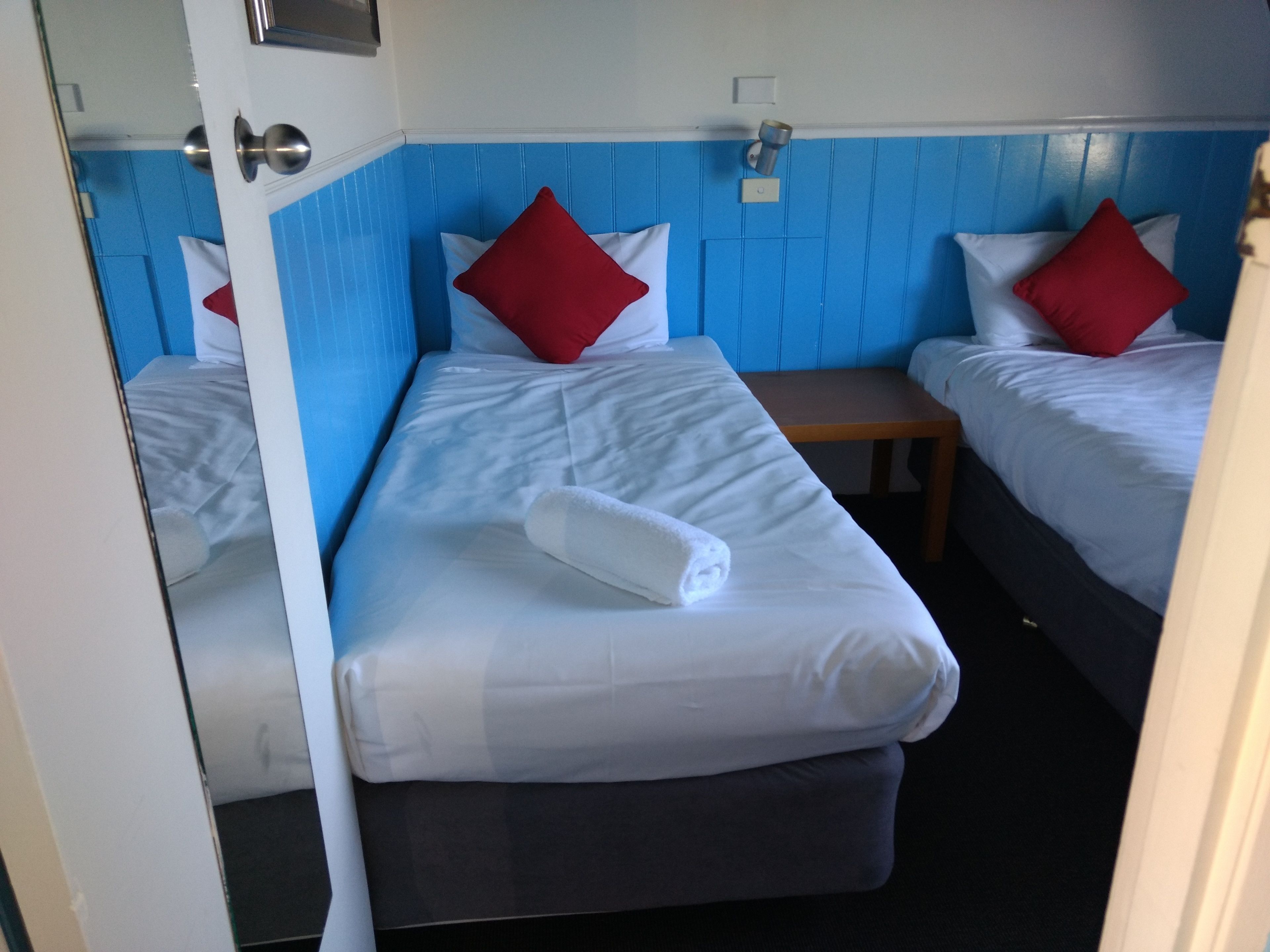 Une chambre d’hôtel avec deux lits, chacun étant doté de draps blancs et d’oreillers rouges. Une table de chevet en bois se trouve entre les deux lits.