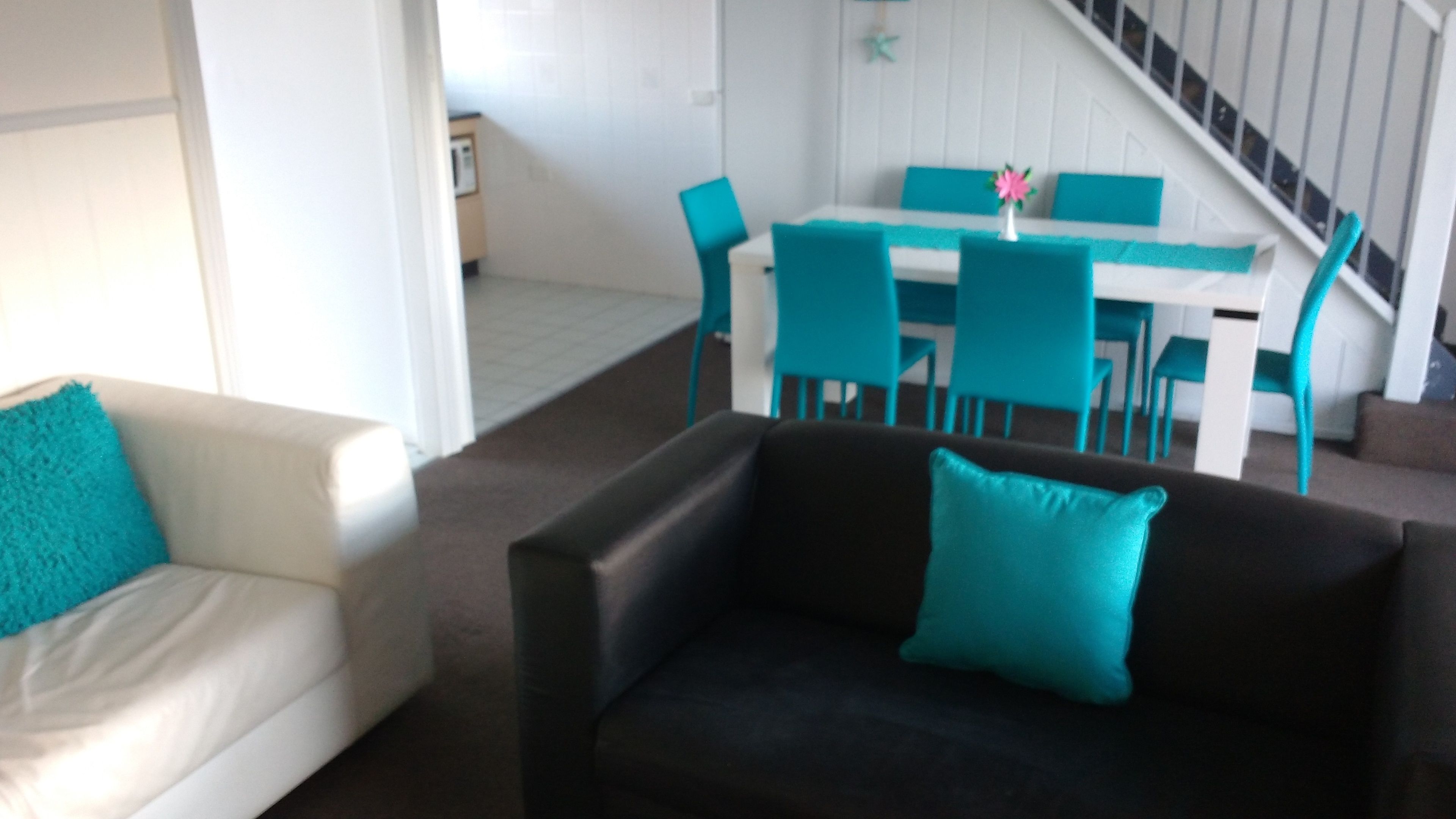 Una sala de estar con un sofá blanco y un sofá negro, ambos con cojines color turquesa. Se observa un comedor con una mesa blanca y sillas color turquesa al fondo.