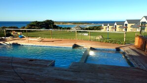 Une piscine avec une terrasse en bois, une vue sur la mer et une pelouse délimitée par une clôture.