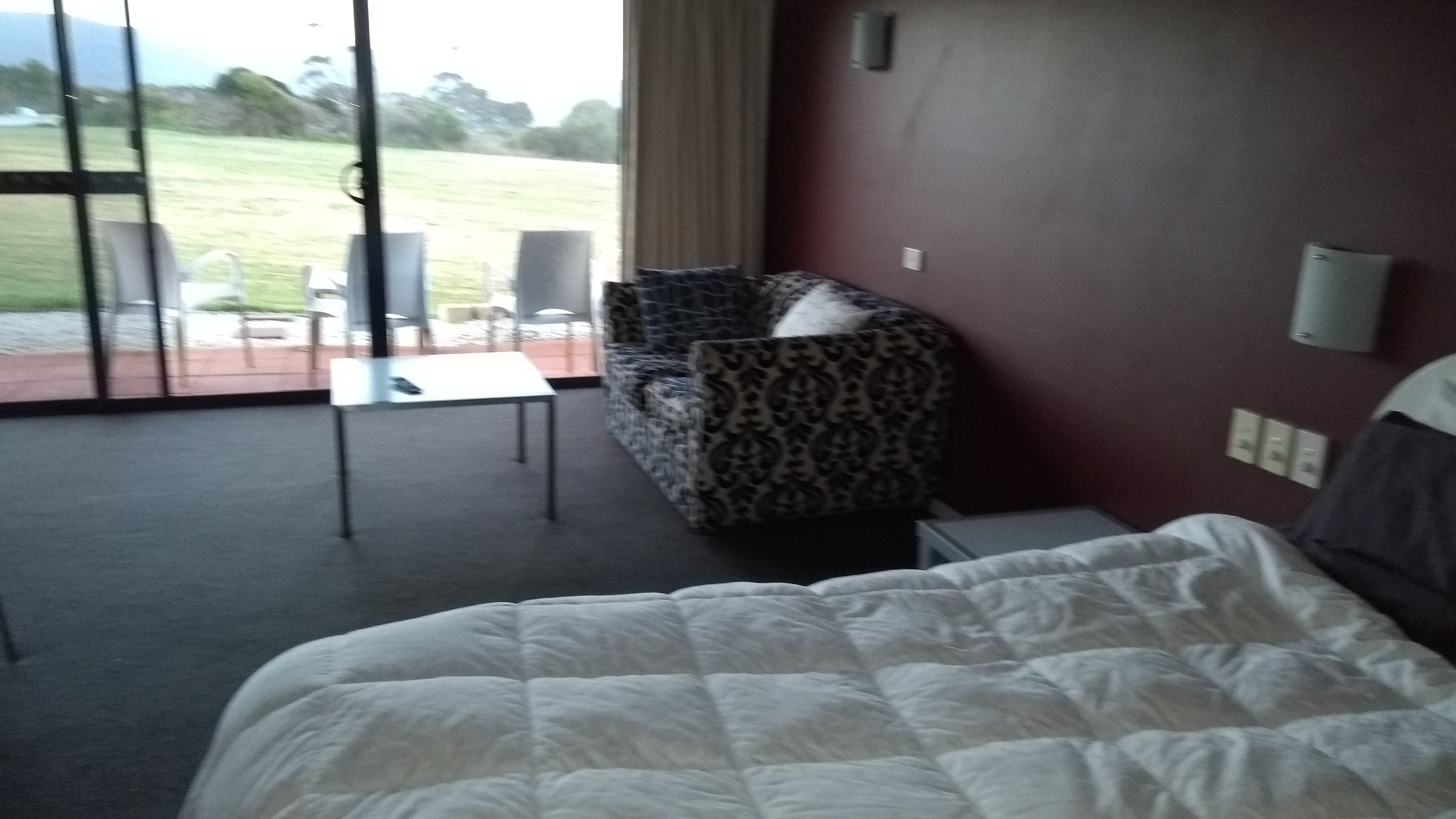 A hotel room with a bed, a patterned armchair, a small table, and a view of a grassy area through large windows.