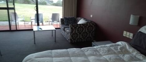 A hotel room with a bed, a patterned armchair, a small table, and a view of a grassy area through large windows.