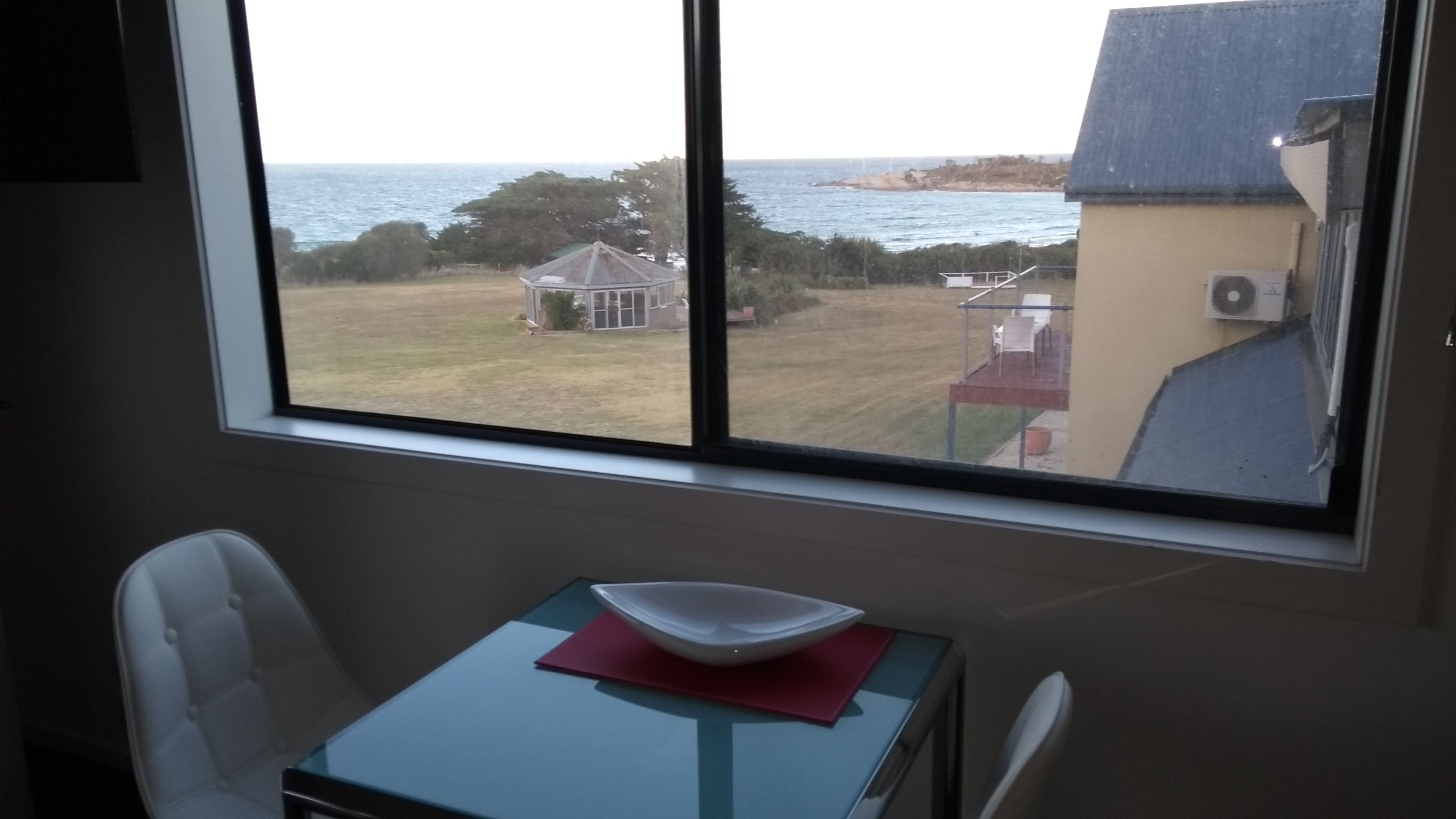 A room with a large window overlooking a grassy area and the sea. There is a table with a white bowl and a red napkin on it.
