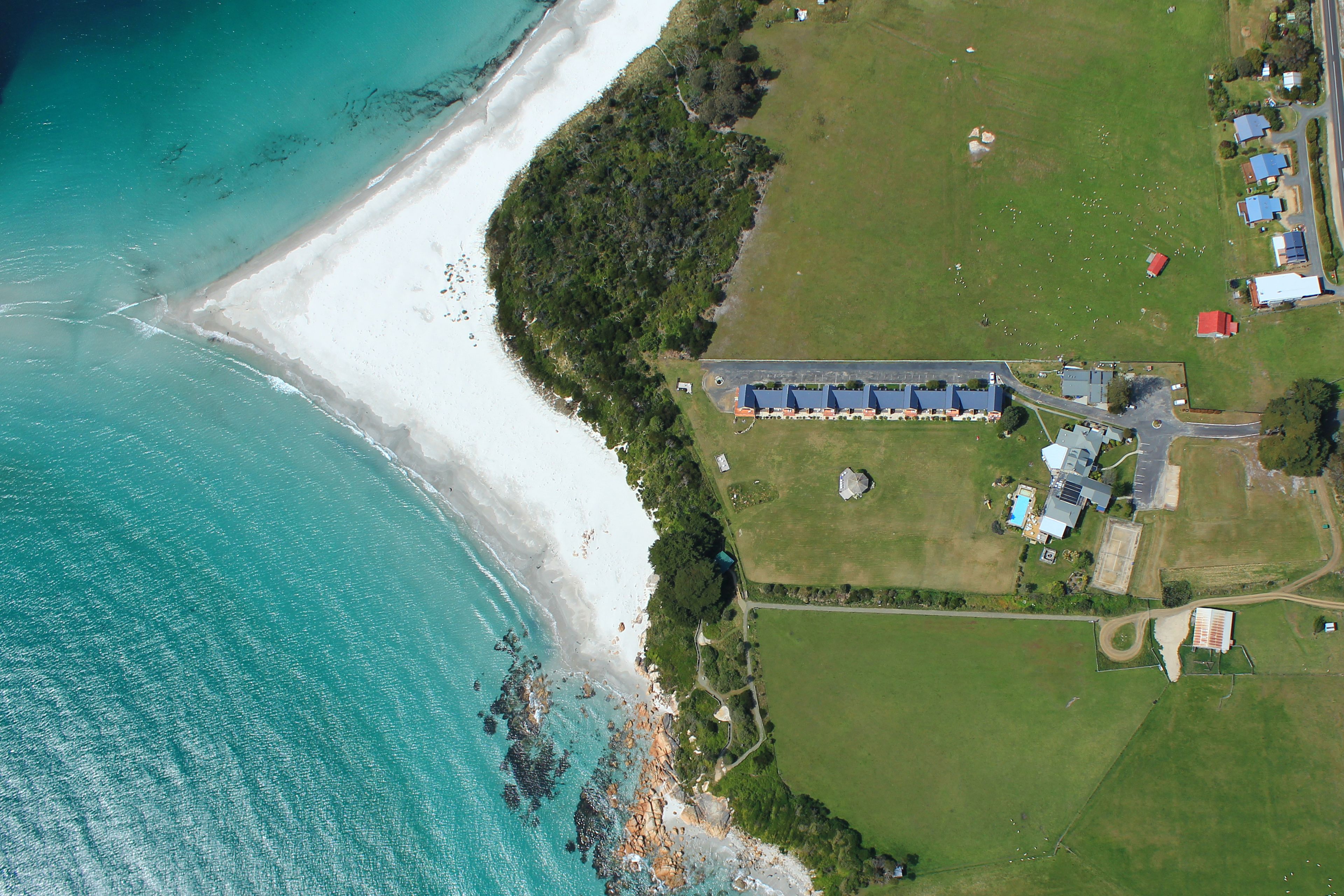 An aerial view of a coastal property with a beach, green fields, and a building complex.