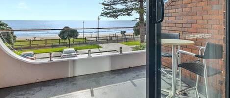 Un balcón con vista a la playa y el océano, una mesa con sillas y un muro de ladrillo.
