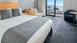 Habitación de hotel con una cama grande, un sofá, una silla, una mesita y un balcón con vistas al mar.