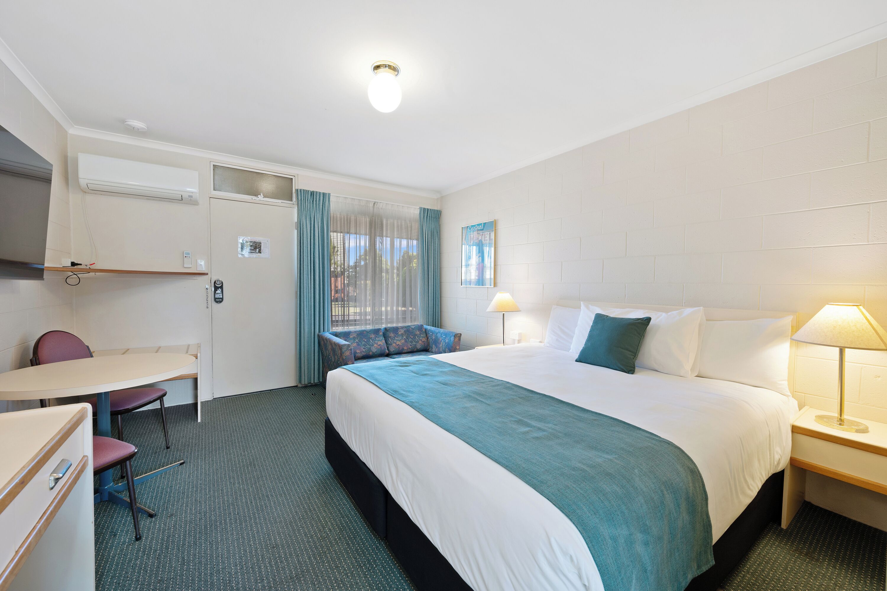 A hotel room with a bed, a desk, a chair, an air conditioning unit, and a window with curtains.