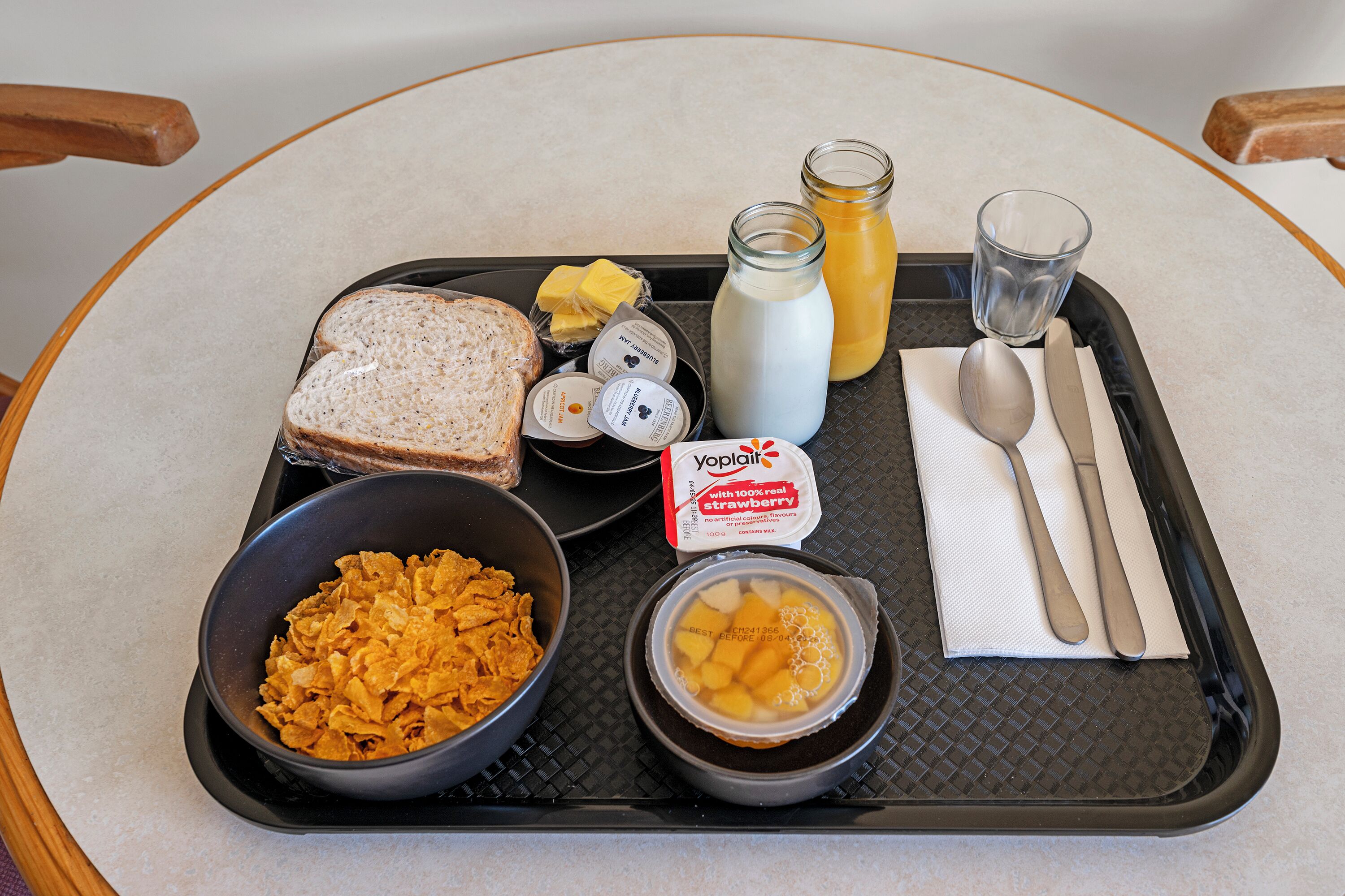 Un plateau contenant des aliments pour le petit-déjeuner, notamment des céréales, des toasts, du beurre, du lait, un verre de jus de fruits et un bol de fruits.