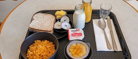 Un plateau contenant des aliments pour le petit-déjeuner, notamment des céréales, des toasts, du beurre, du lait, un verre de jus de fruits et un bol de fruits.