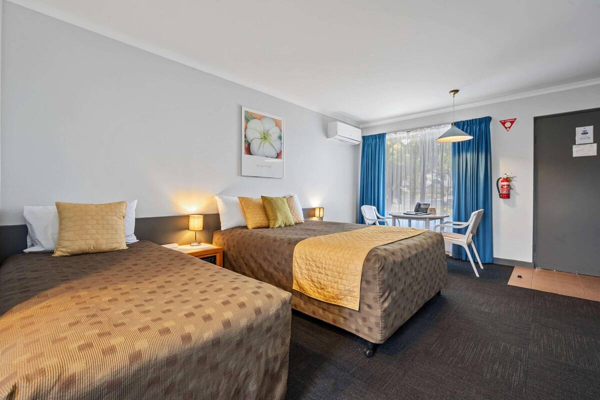 A hotel room with two beds, a desk, a chair, a fire extinguisher, and a window with curtains.