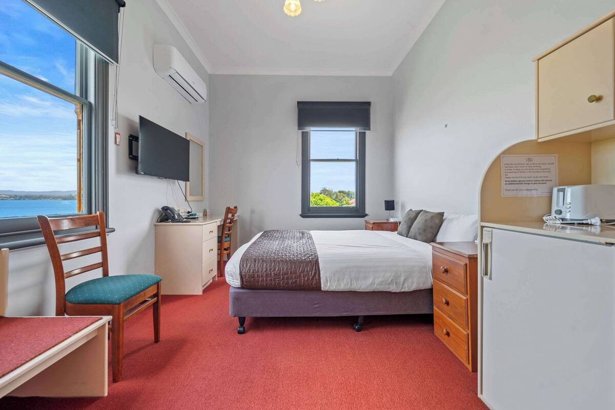 A bedroom with a bed, a desk, a chair, a TV, a window with a view, and a refrigerator.