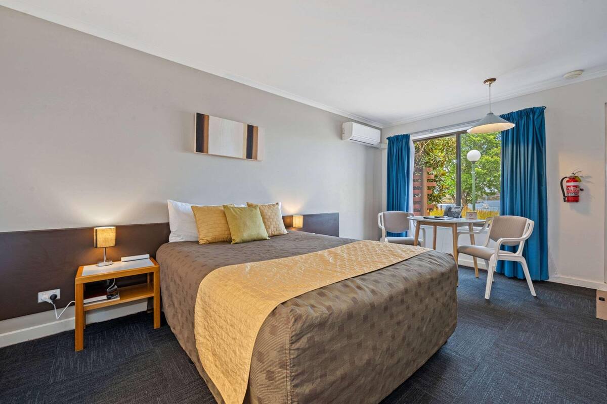 A hotel room with a large bed, bedside table with a lamp, a small table with chairs, and a view of the outdoors through large windows.