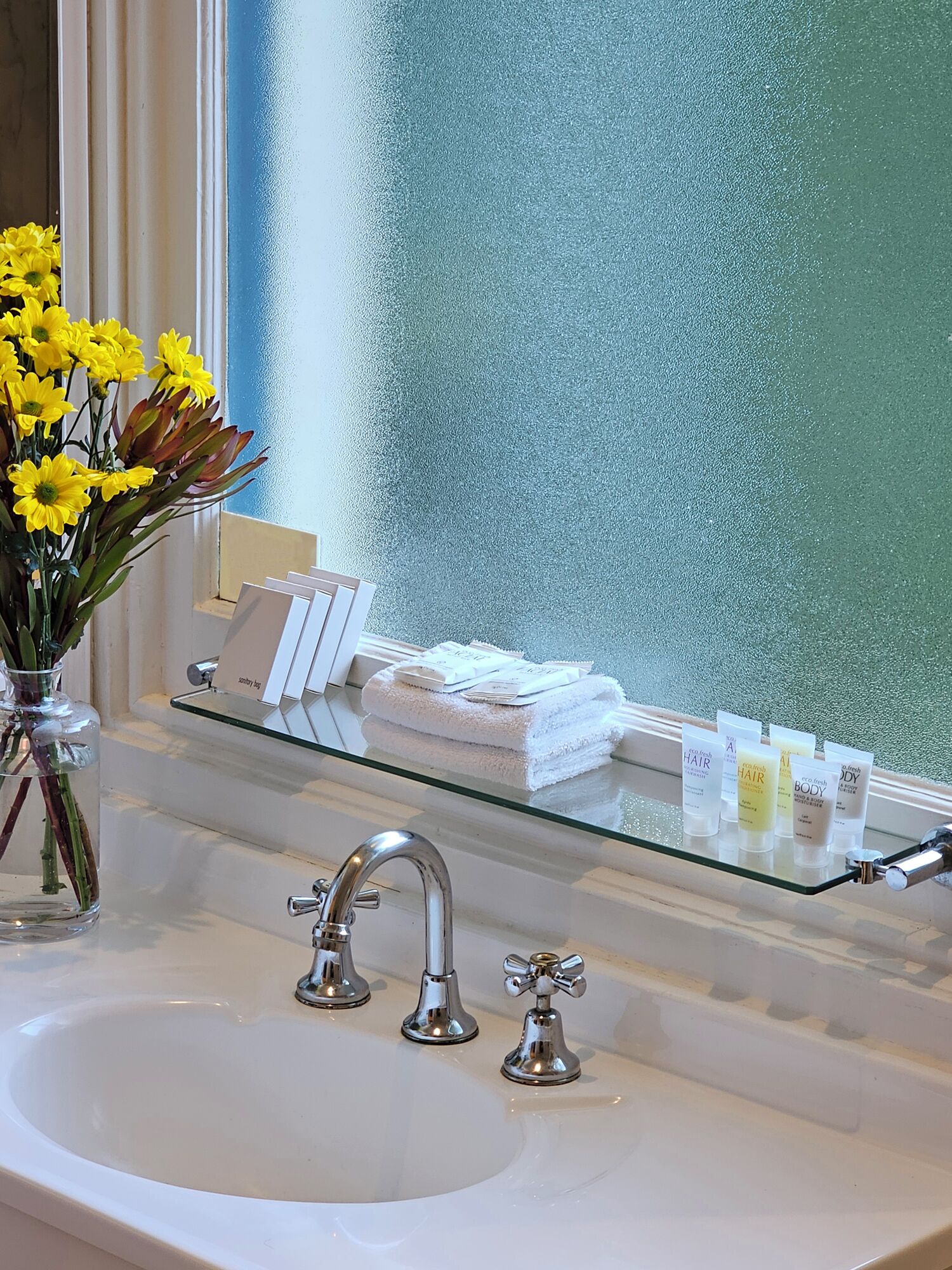 A bathroom with a glass shelf holding toiletries, a vase with yellow flowers, and a window with condensation.
