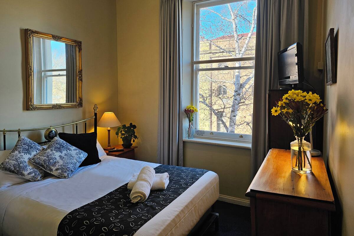 A hotel room with a bed, a bedside table with a lamp, a mirror, a window with curtains, and a vase with flowers.