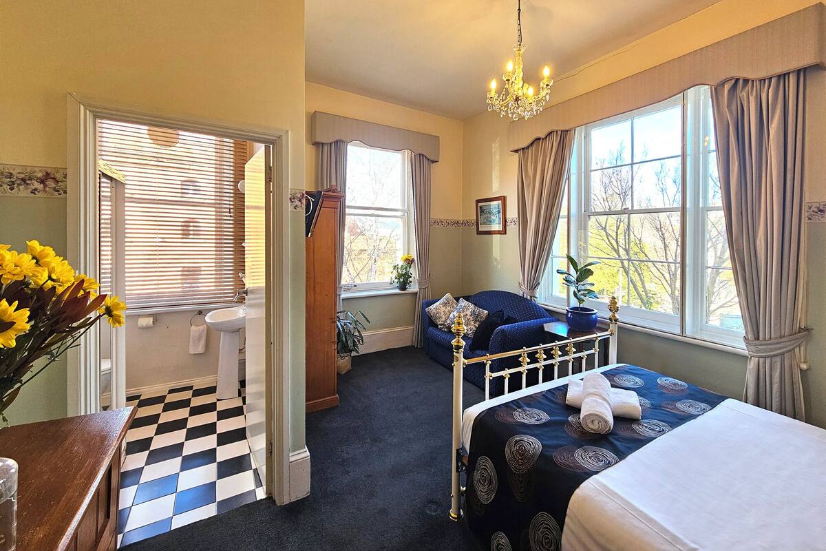 A bedroom with a checkered floor, a bed with a blue and white patterned bedspread, a chandelier, and a window with curtains.