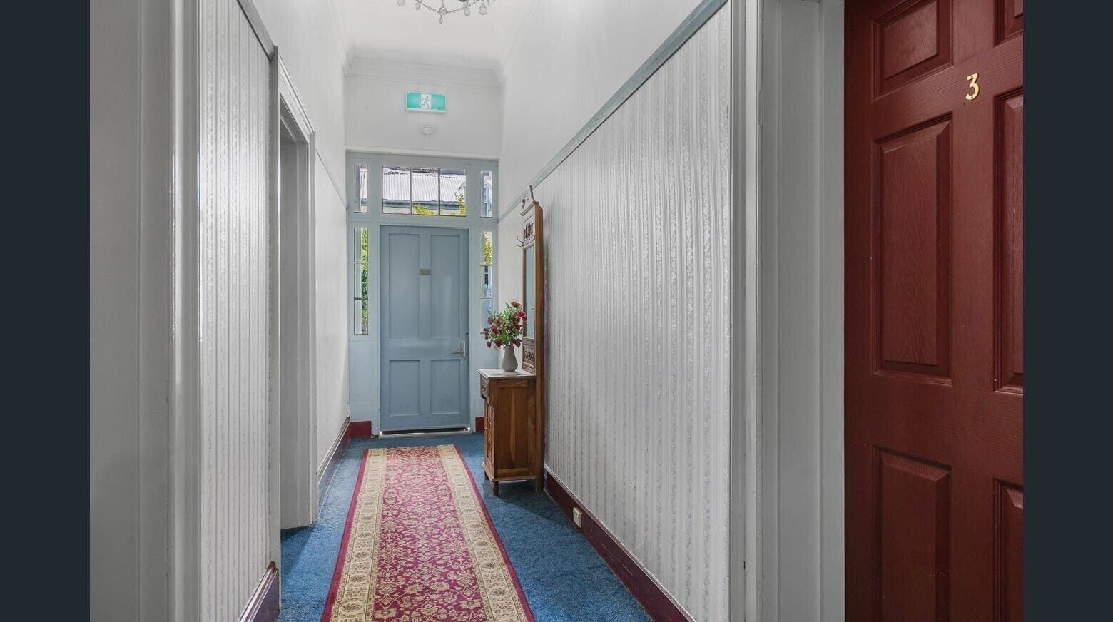 A hallway with a blue carpet, a wooden cabinet with a vase of flowers, and a red door with the number 3.