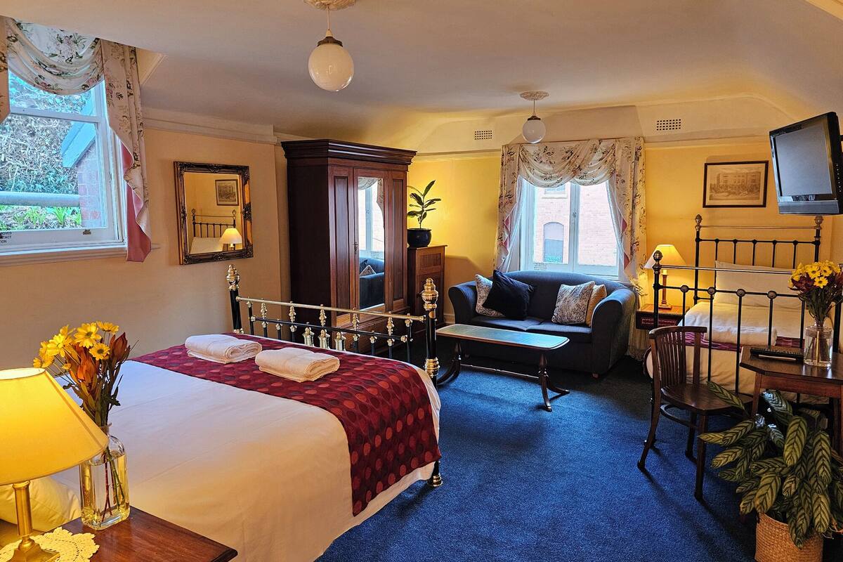 A hotel room with a bed, a sofa, a TV, and a desk with a lamp and flowers.
