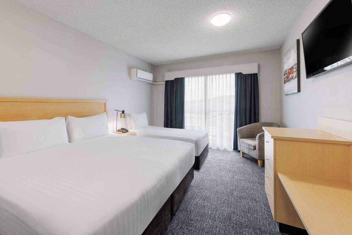 Quarto de hotel com duas camas, uma escrivaninha, uma cadeira, uma televisão e uma janela com cortinas.