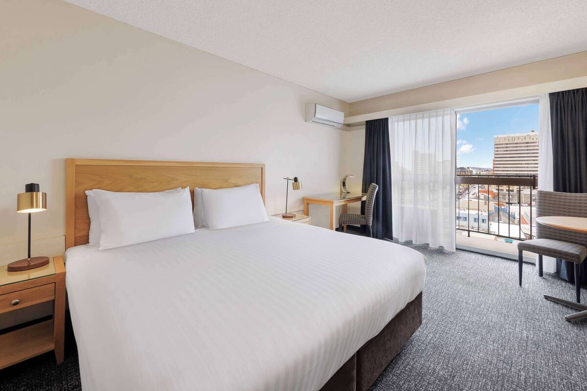 Quarto de hotel com uma cama grande, uma escrivaninha, uma cadeira e uma varanda com vista para a cidade.