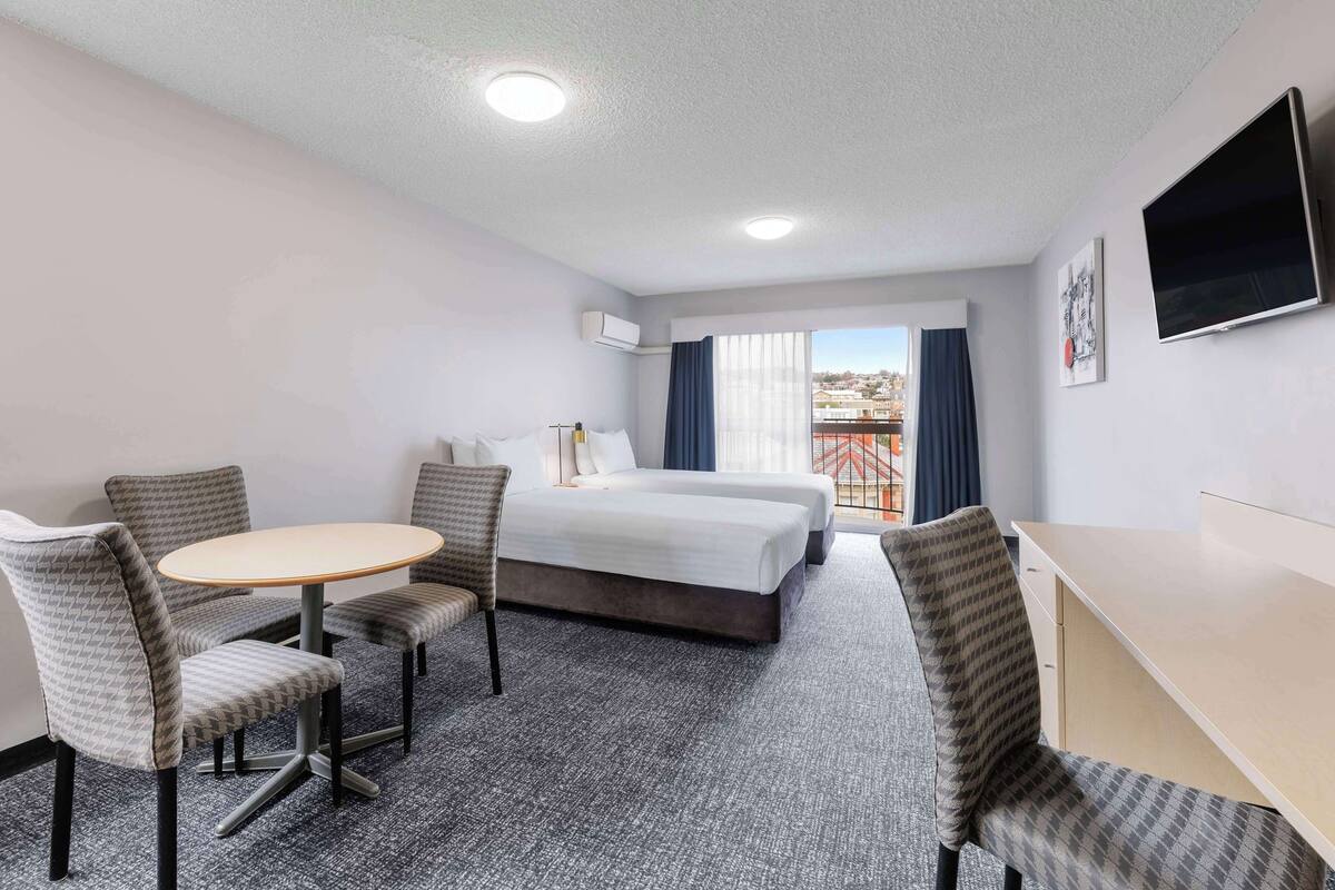 Quarto de hotel com uma cama, duas cadeiras, uma mesa pequena, escrivaninha, televisão e janela com cortinas.