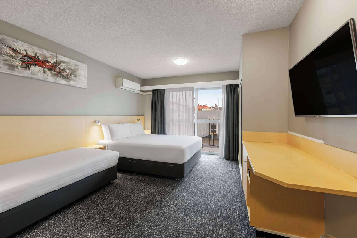 Quarto de hotel com duas camas, uma escrivaninha, televisão de tela plana e uma janela grande com cortinas.