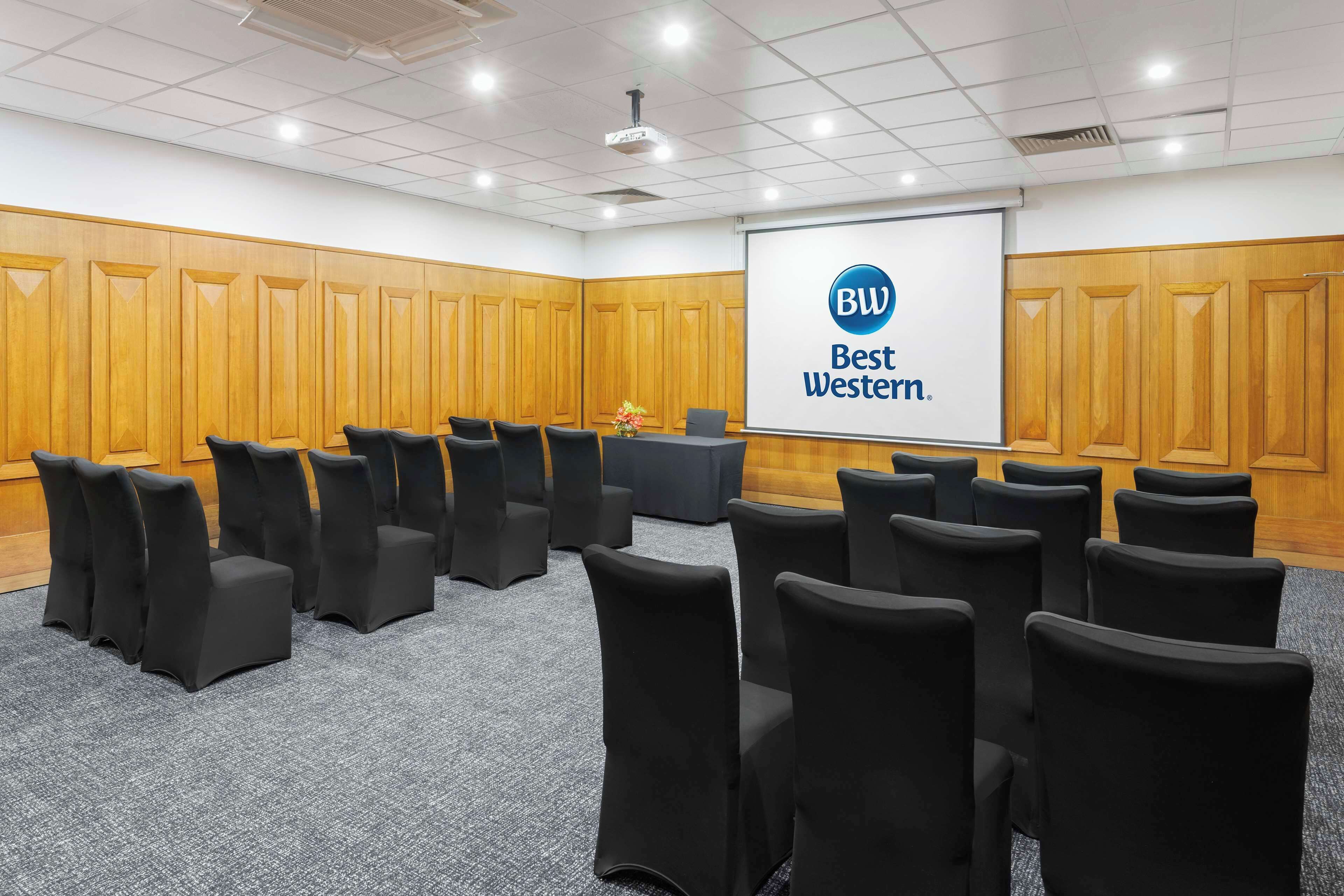 A conference room with rows of black chairs, a projector screen displaying "BW Best Western," and wooden paneling on the walls.