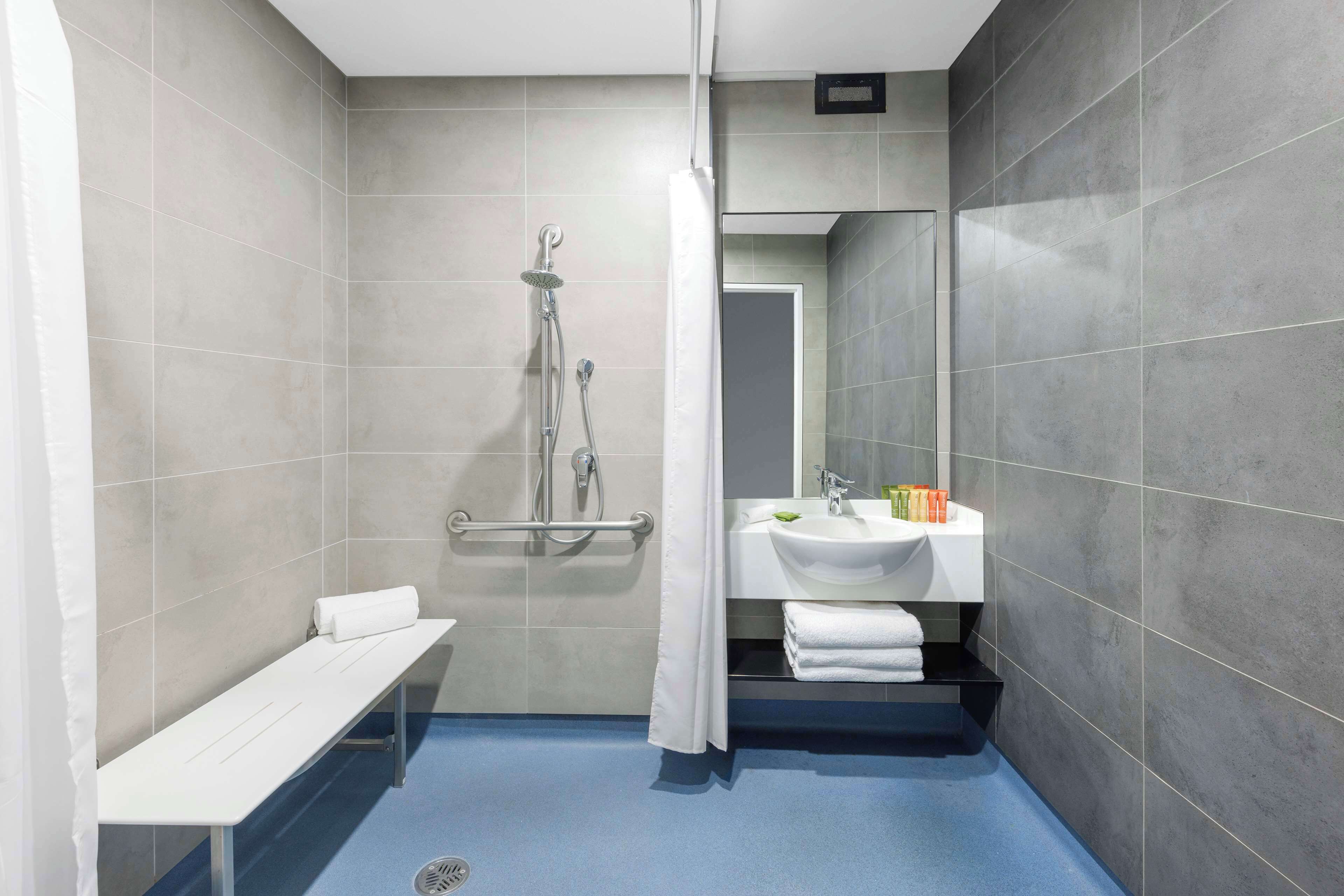 A modern bathroom with grey tiles, a shower with a handheld showerhead, a bench, and a sink with toiletries.