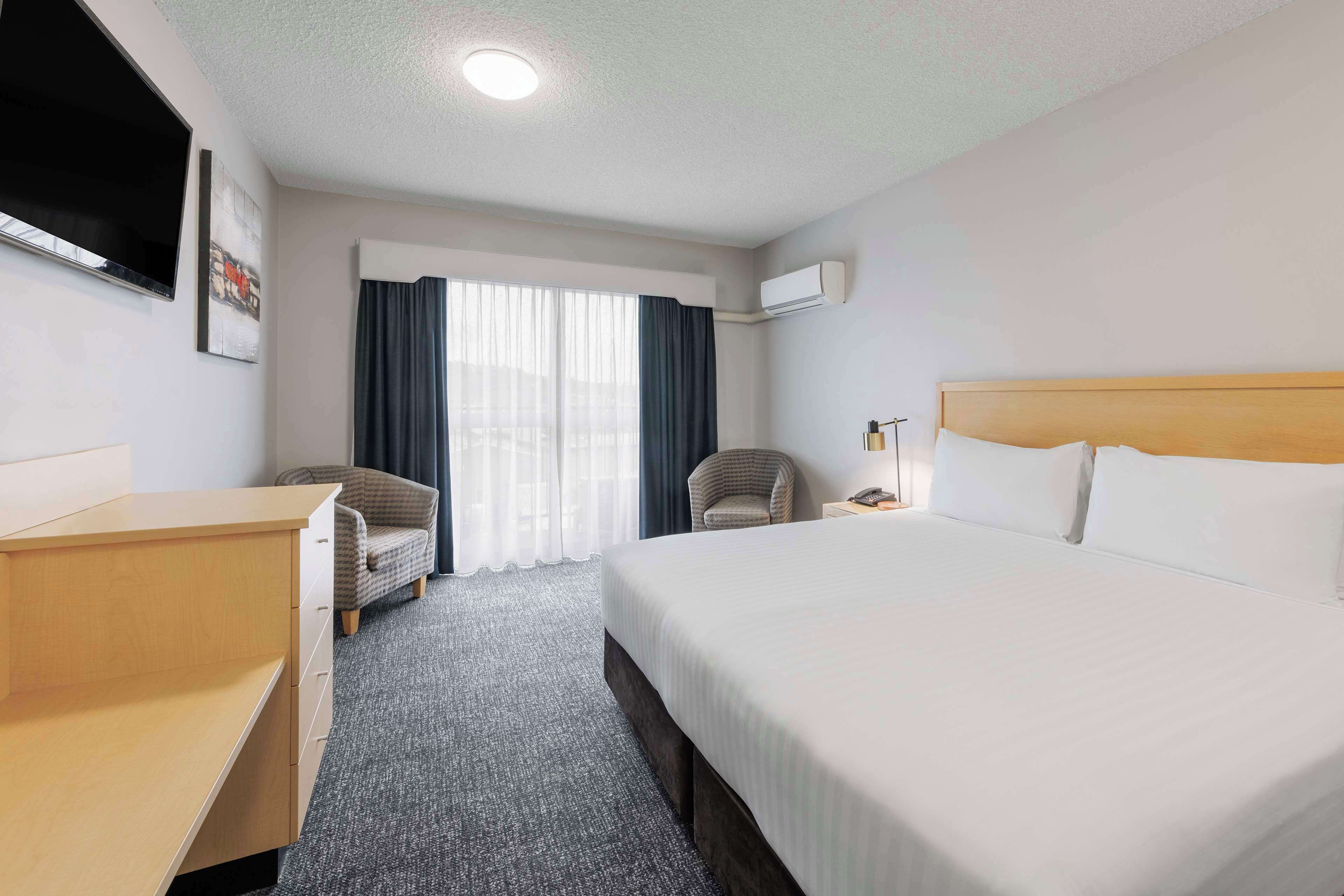 A hotel room with a bed, a desk, a chair, a TV, and an air conditioning unit.