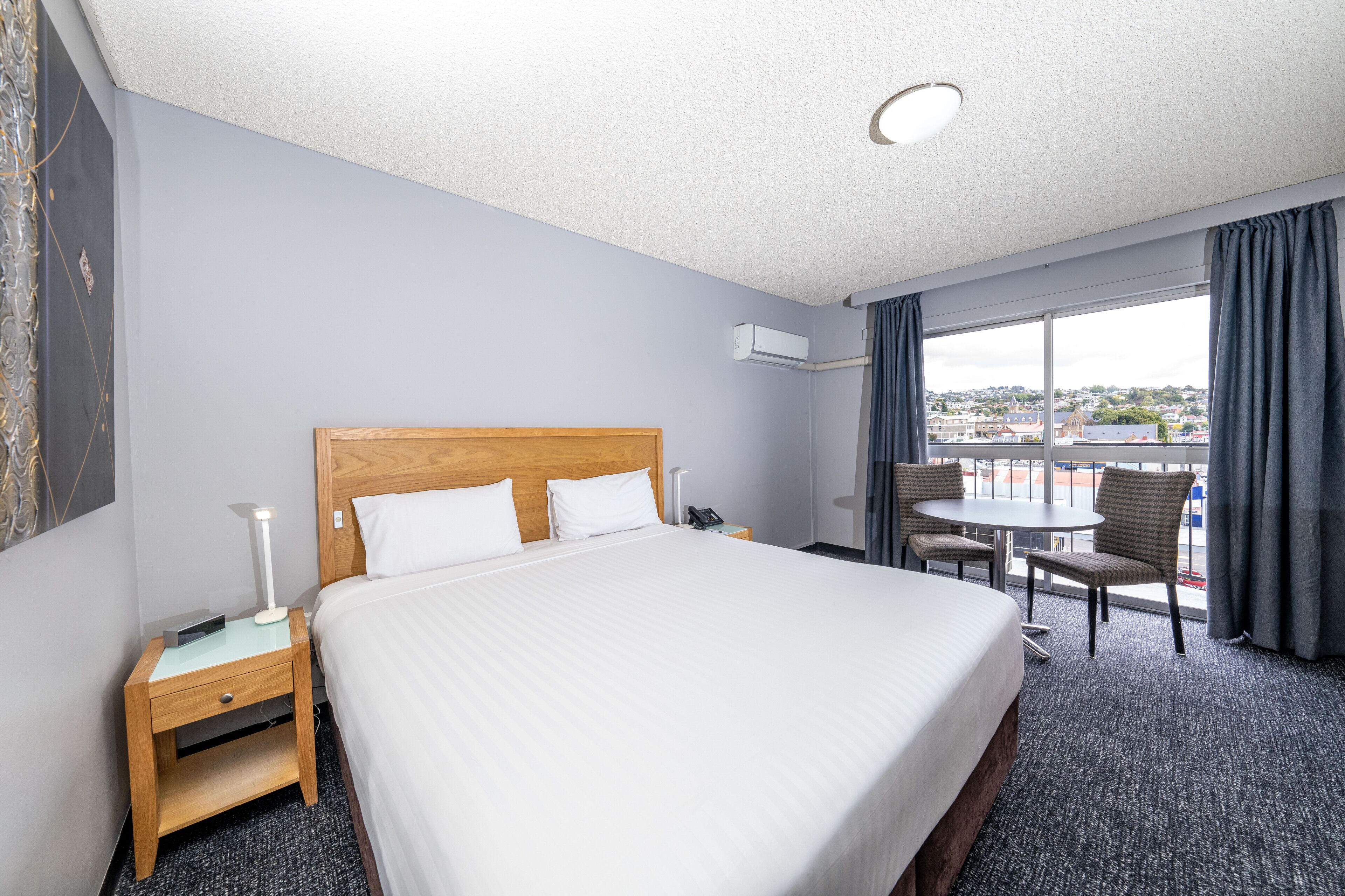 A hotel room with a large bed, bedside tables, a desk with a chair, and a view of the city through large windows.