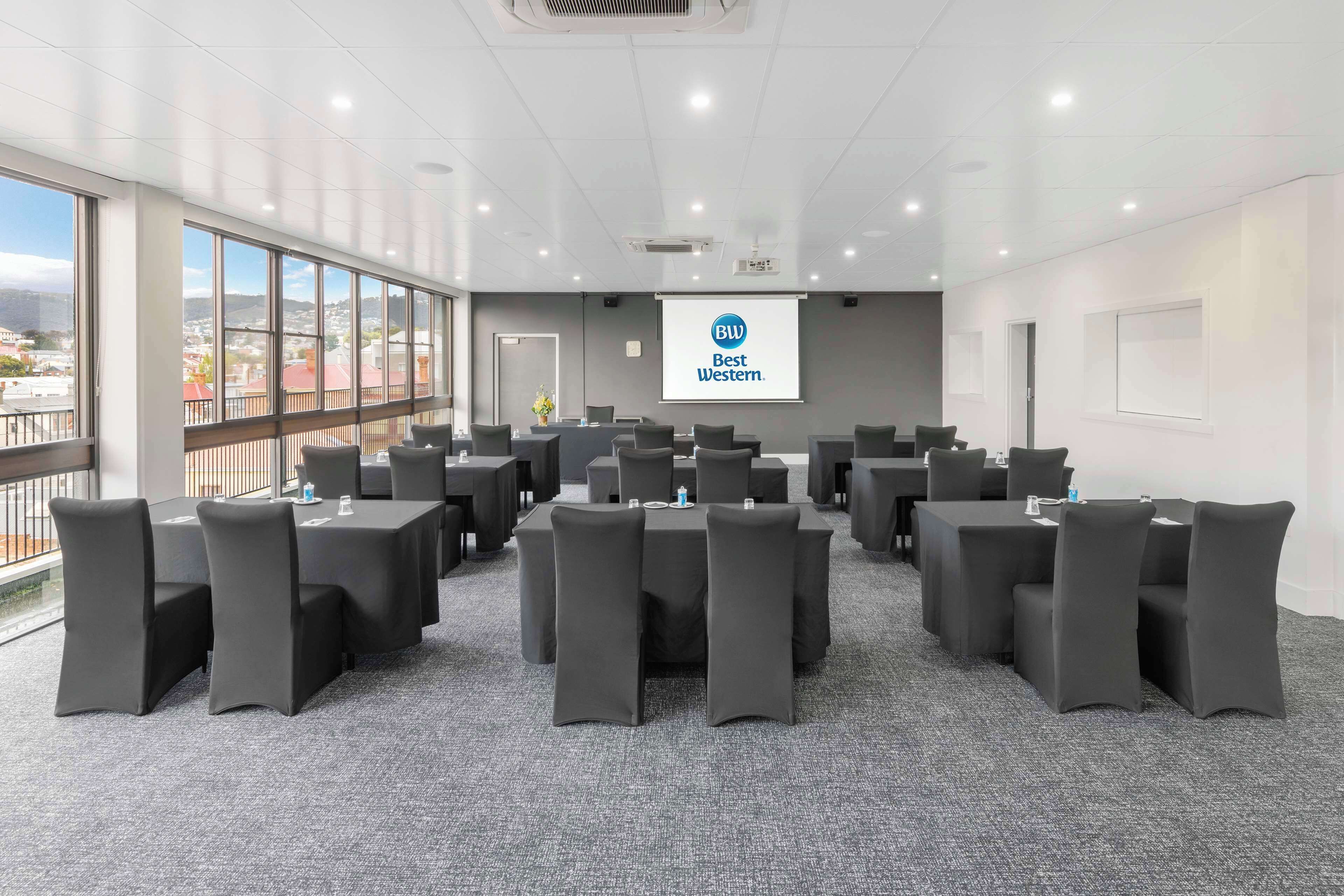 Un salón de conferencias con una pantalla grande mostrando el logotipo de Best Western, varias mesas con sillas y vistas a edificios a través de amplias ventanas.