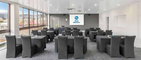 Un salón de conferencias con una pantalla grande mostrando el logotipo de Best Western, varias mesas con sillas y vistas a edificios a través de amplias ventanas.