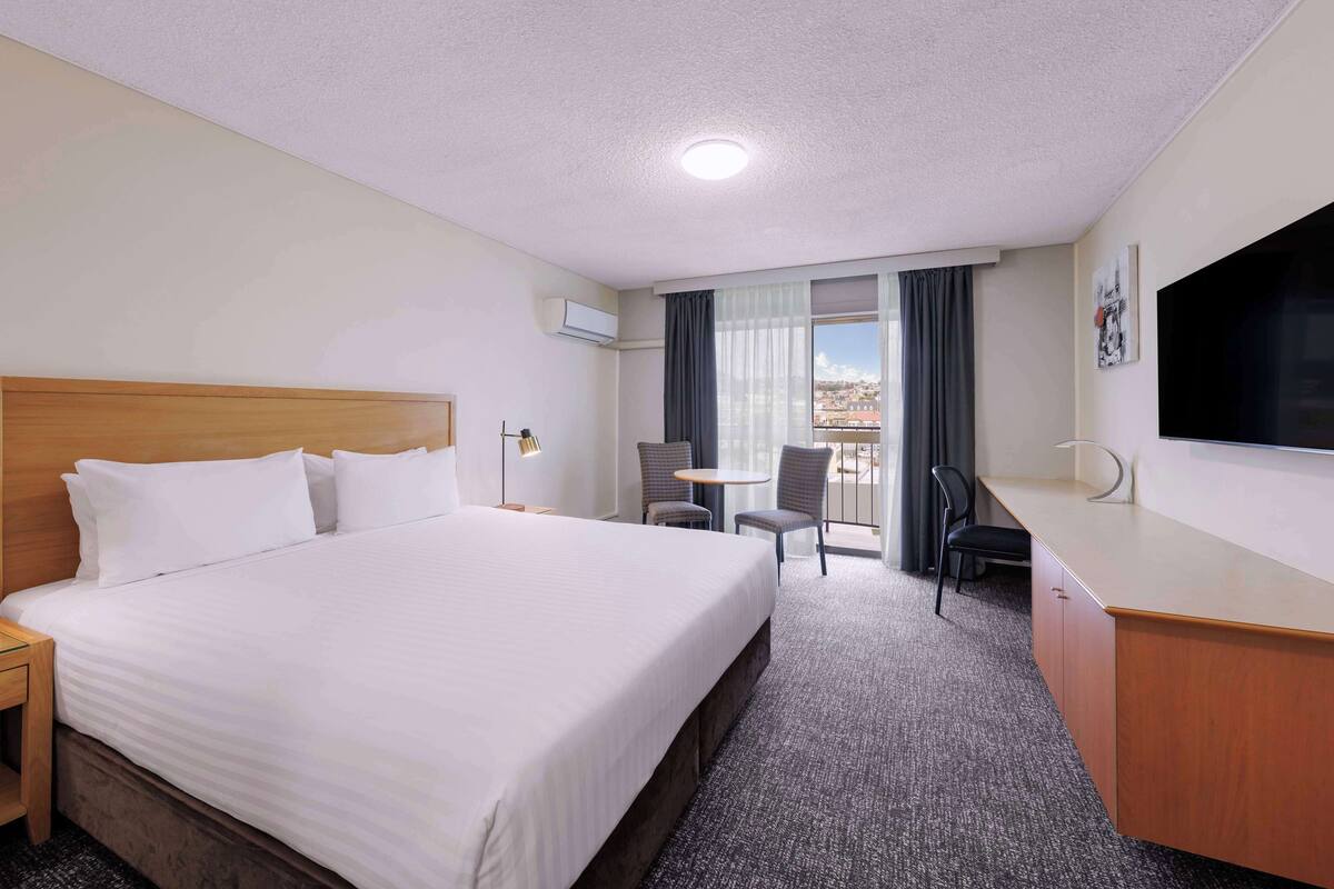 Quarto de hotel com uma cama grande, escrivaninha, cadeira, televisão e varanda com cortinas.