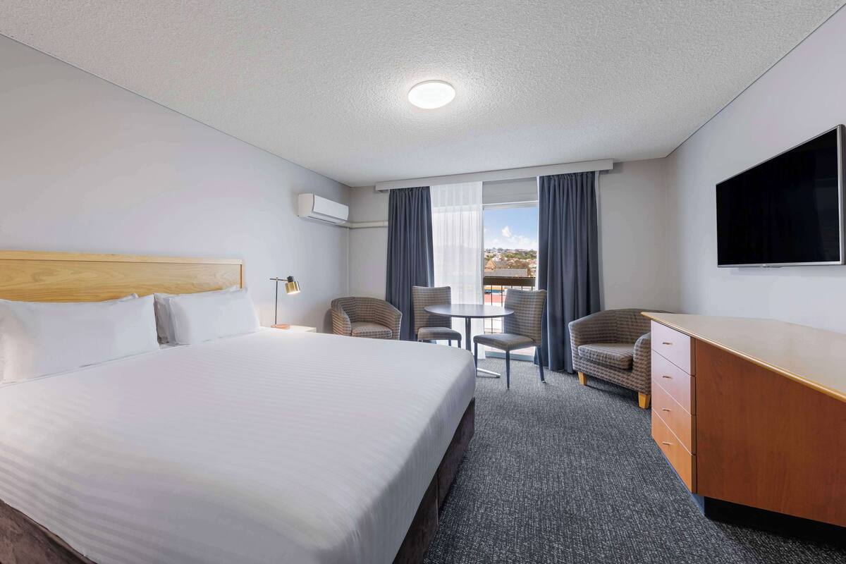 Quarto de hotel com uma cama grande, uma escrivaninha, duas cadeiras, uma televisão e uma janela com cortinas.