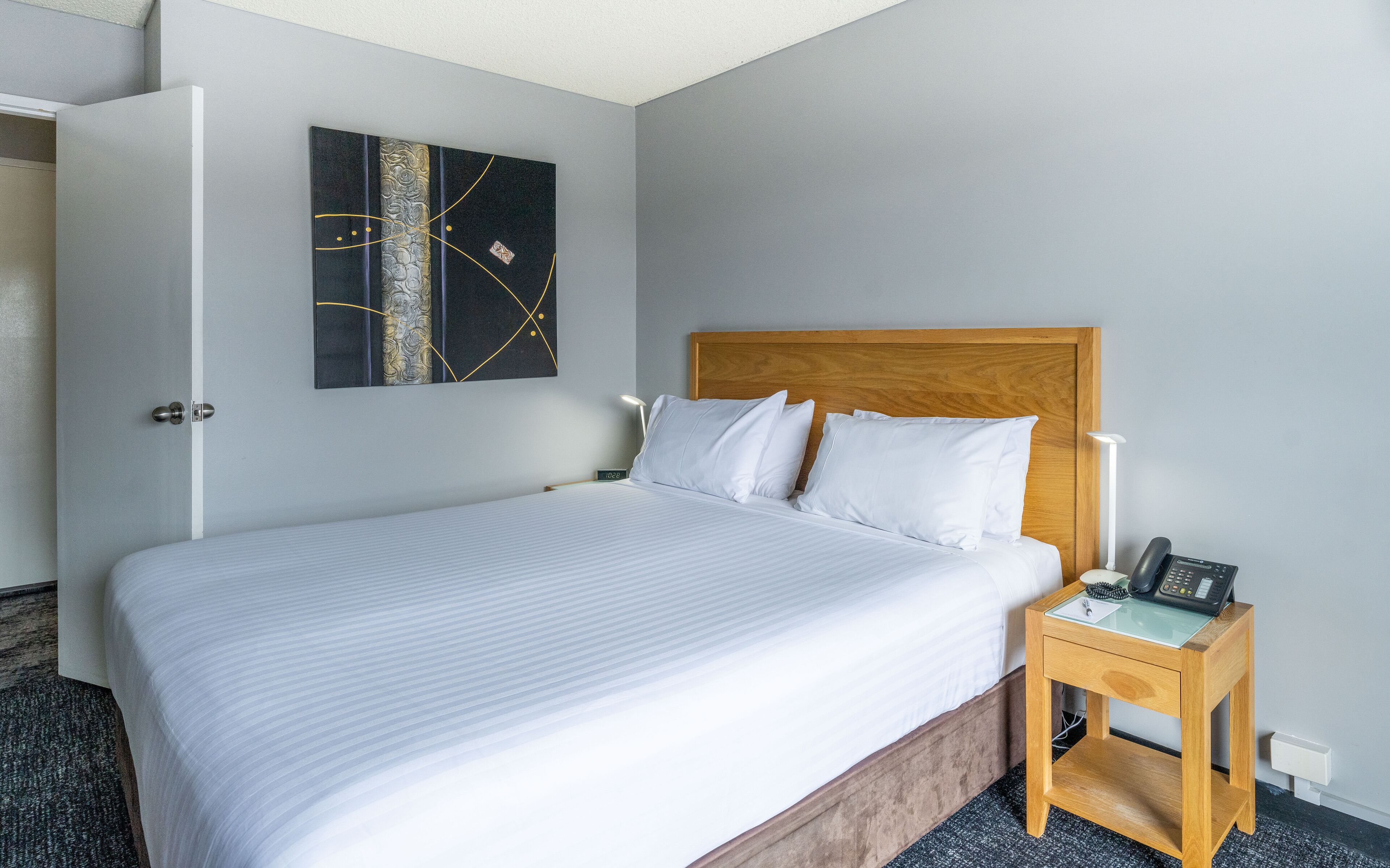 A hotel room with a large bed, a wooden headboard, a bedside table with a lamp, and a wall-mounted artwork.
