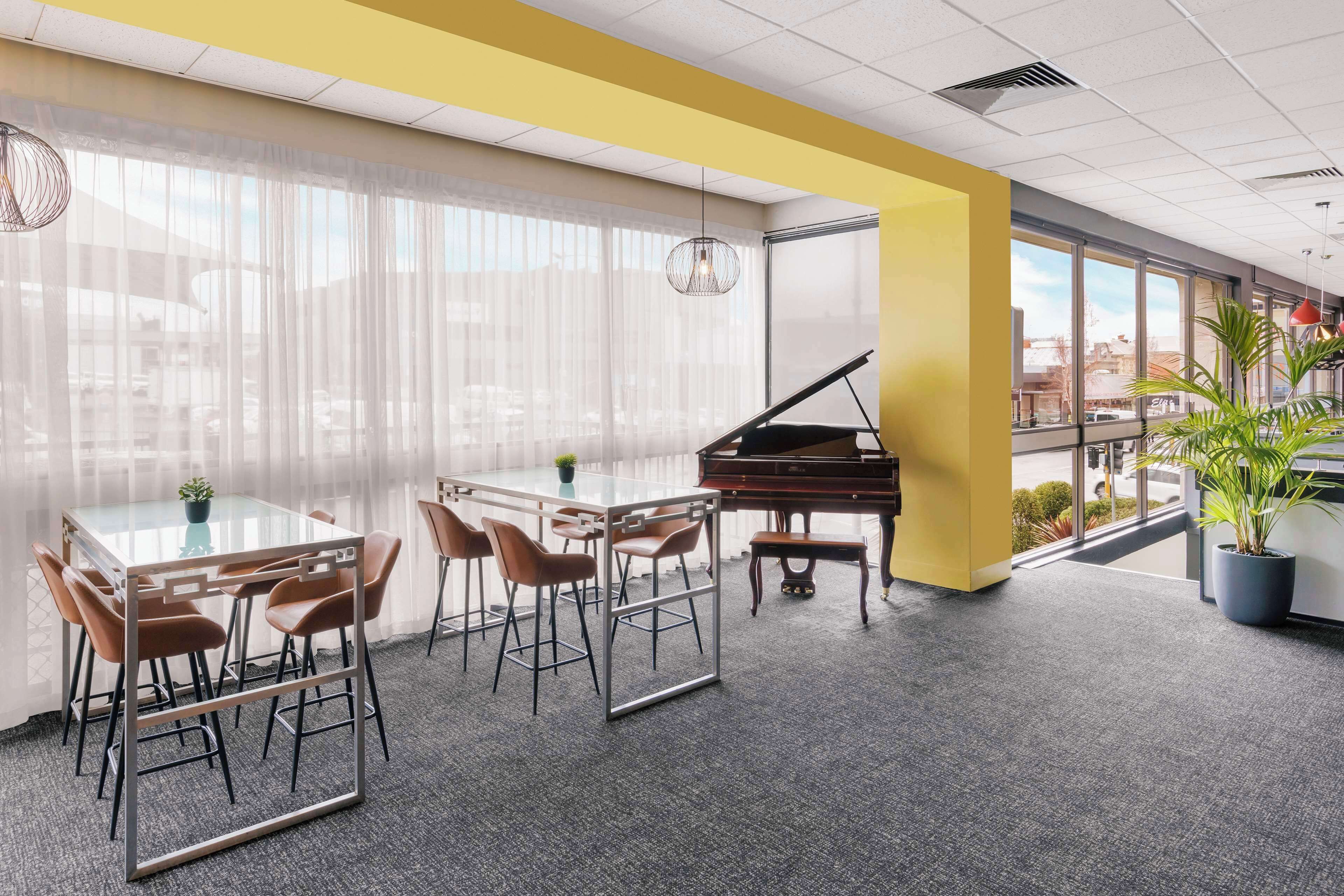 A modern conference room with a glass table, brown chairs, a grand piano, and large windows with sheer curtains.