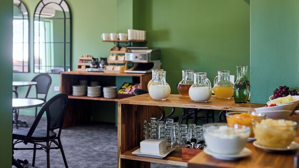 A breakfast setup with a variety of food items and beverages on a wooden table, surrounded by chairs and a green wall. - Balmoral On York (Launceston)