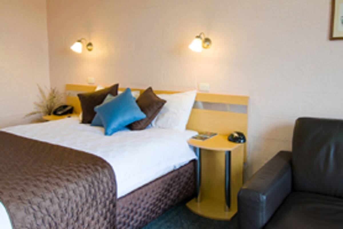 A hotel room with a bed, a bedside table with a lamp, a chair, and a wall-mounted phone.