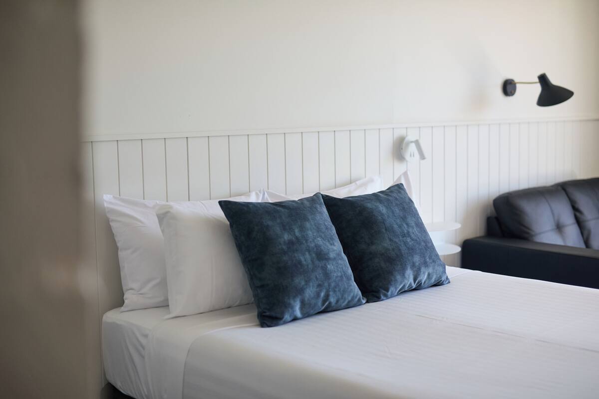 A neatly made bed with white and dark blue pillows, a black lamp on the wall, and a dark sofa in the background.