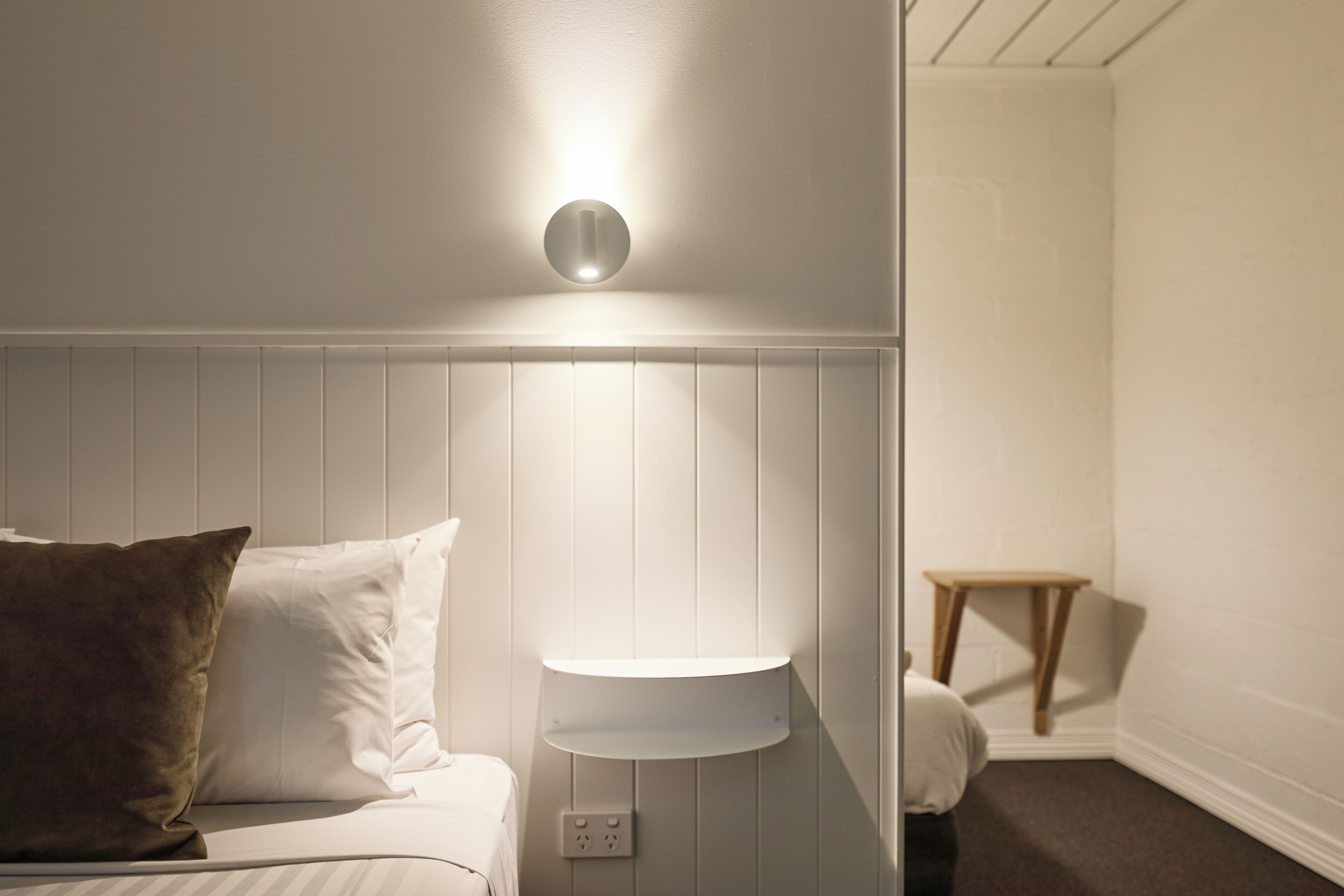 A bedroom with a bed, a bedside table, a wall-mounted light, and a small wooden stool.